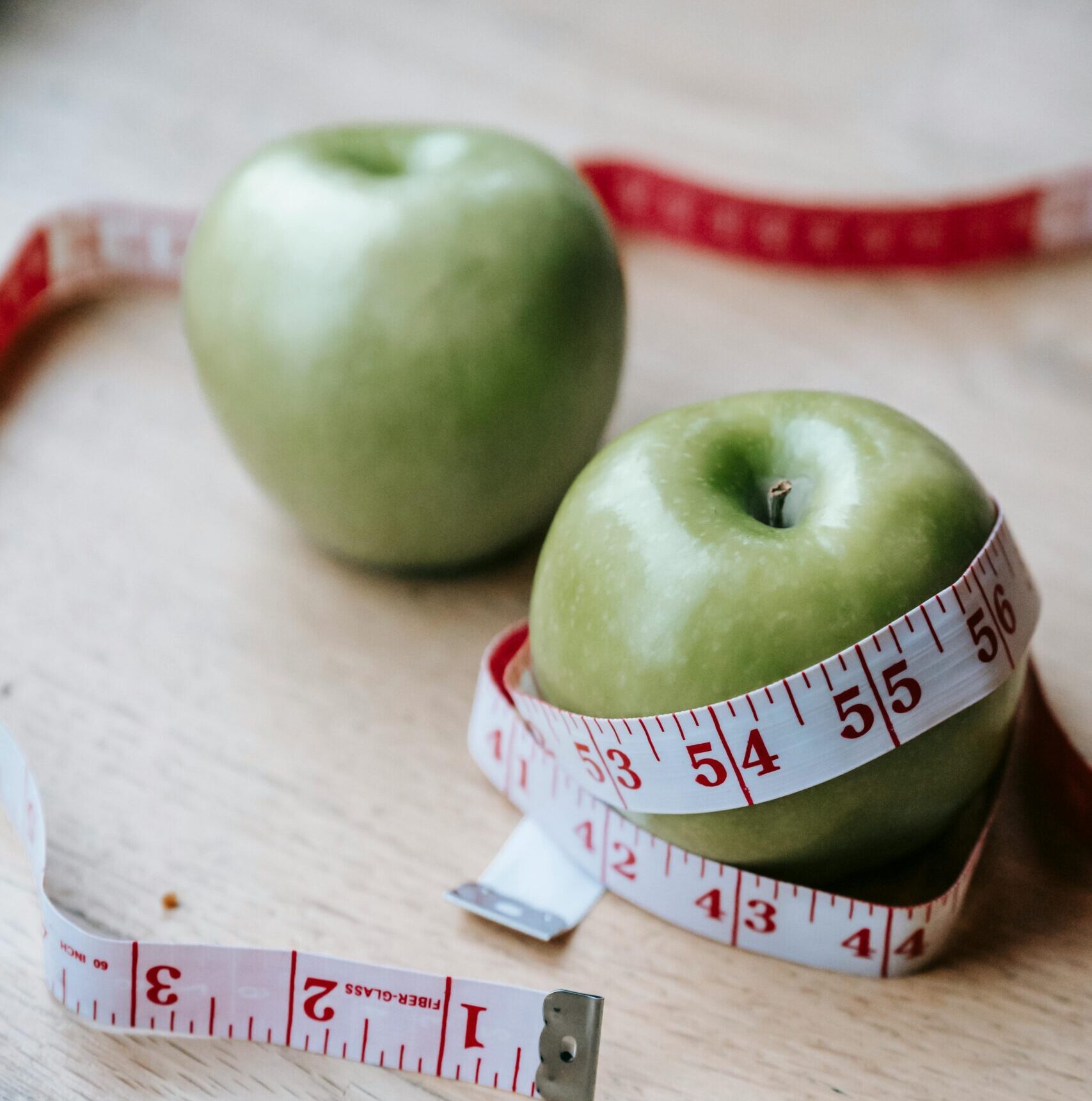 Two green apples on a wooden surface wrapped with a red and white measuring tape, symbolizing health and diet.